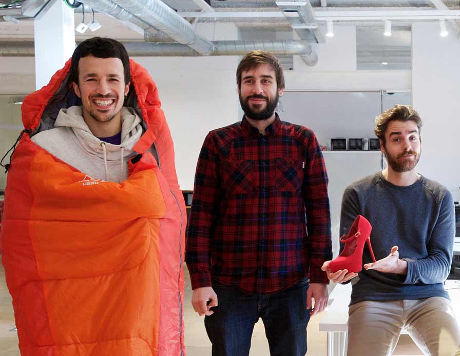 A man in a sleeping bag stands next to a man in a plaid flannel next to a man holding a red high heel